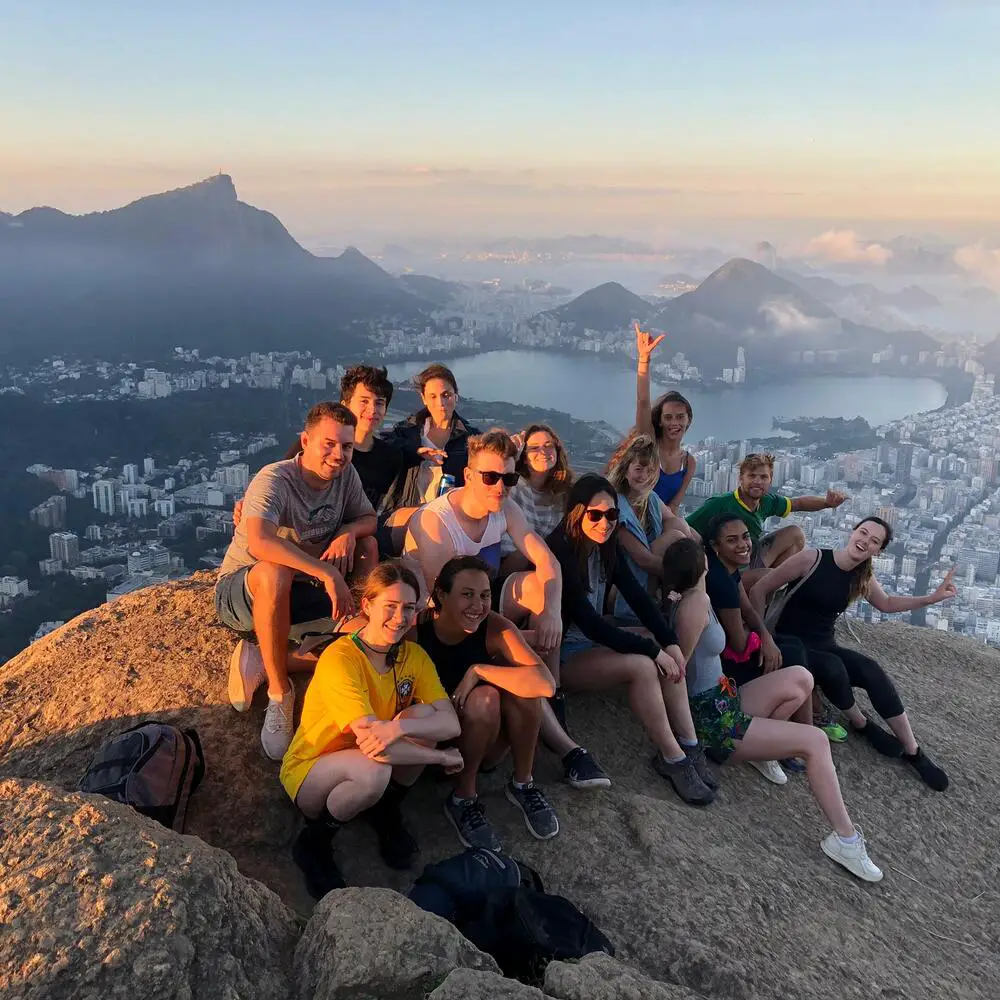 Hikers enjoying a scenic view from the summit of Dois Irmãos mountain, with Rio de Janeiro's skyline and coastline stretching out beneath them, bathed in golden sunlight.
